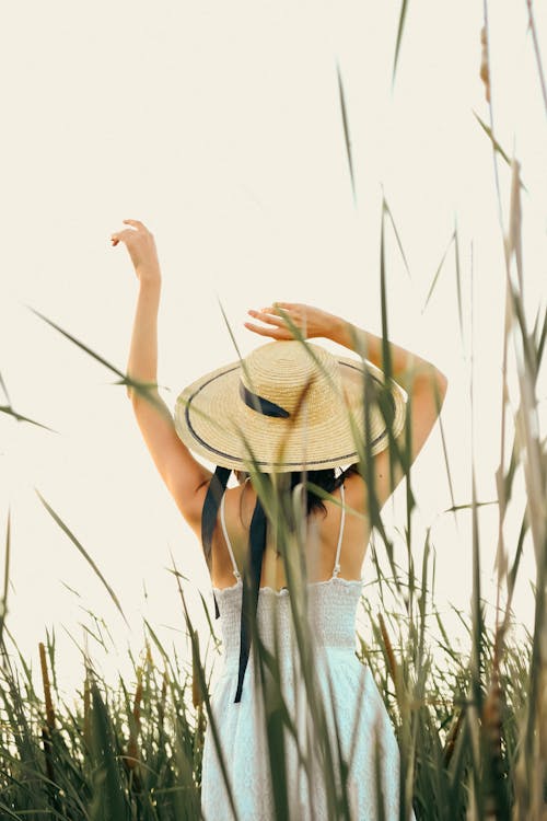 Woman in Hat and Dress Standing with Arms Raised among Grasses 