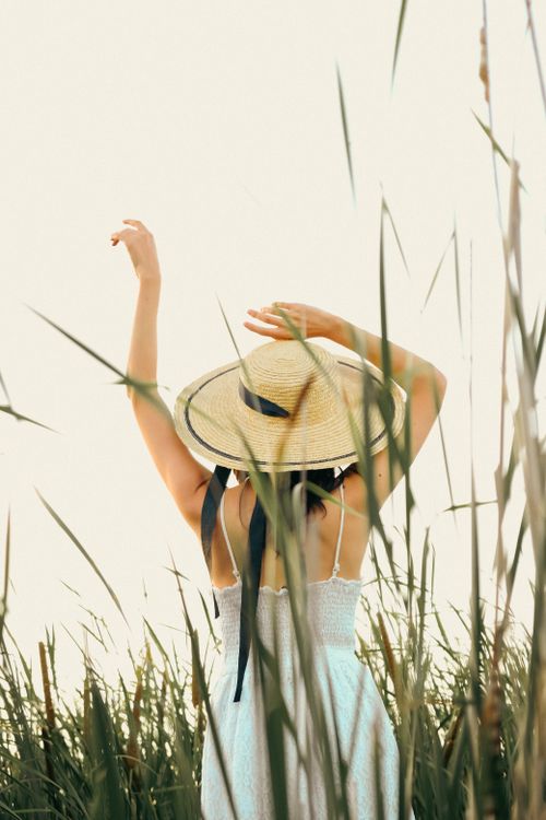Free A woman in a summery dress and straw hat stands among tall grasses in a rural setting. Stock Photo