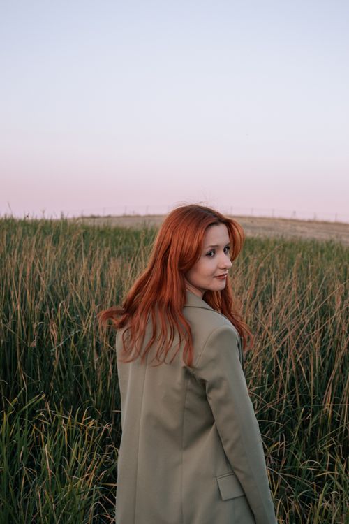 Free Redhead woman stands in a rural field at sunset, looking back with long flowing hair. Stock Photo