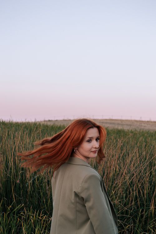 Free A woman with long red hair in a coat stands in a rural field at sunset. Stock Photo