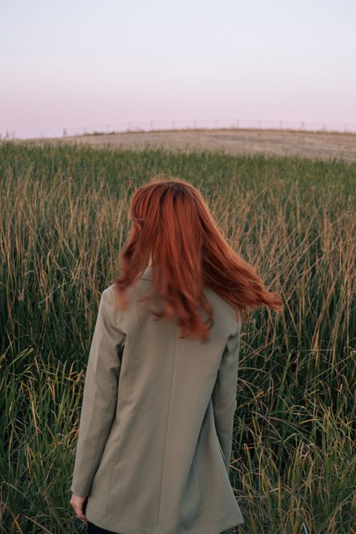 Free A woman with striking red hair stands in a lush green field at sunset. Stock Photo