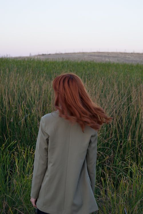 Free A serene scene of a woman with red hair standing in a grassy field at dusk, showcasing fashion and nature Stock Photo