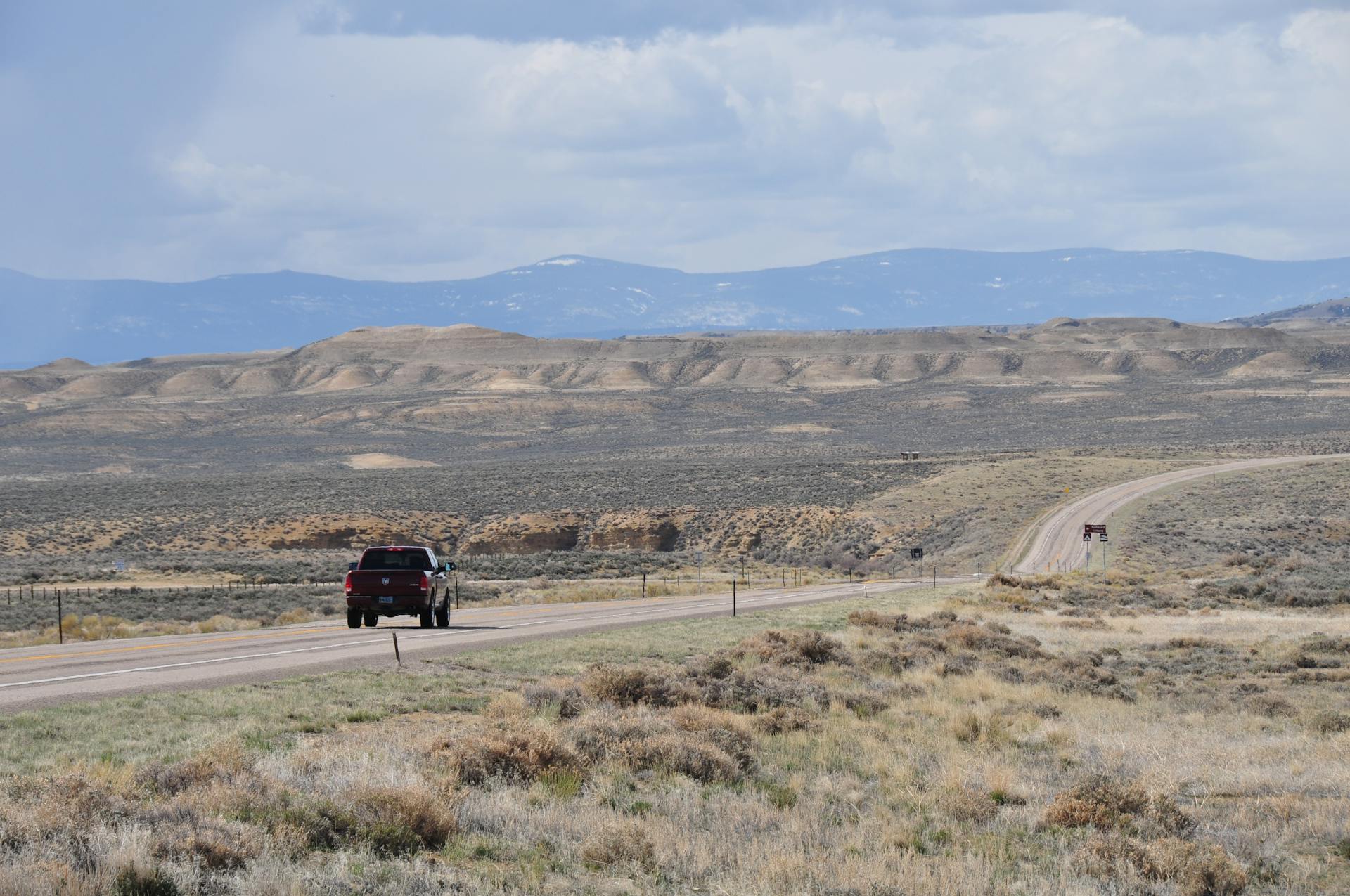 A solitary truck travels along a desert highway with mountainous terrain in the distance.