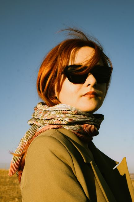 Elegant woman with red hair wearing sunglasses and a scarf in a sunny outdoor setting, exuding fashion and confidence.