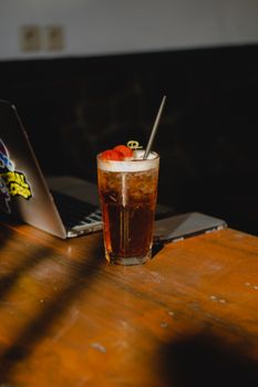 A chilled drink garnished with strawberries next to a laptop on a sunny wooden table.