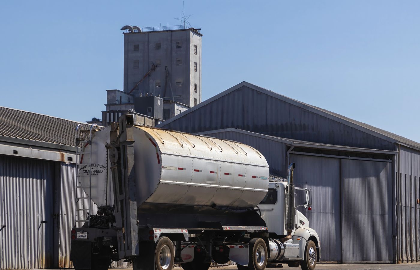 A large semi-trailer truck parked at an industrial grain warehouse under clear skies.