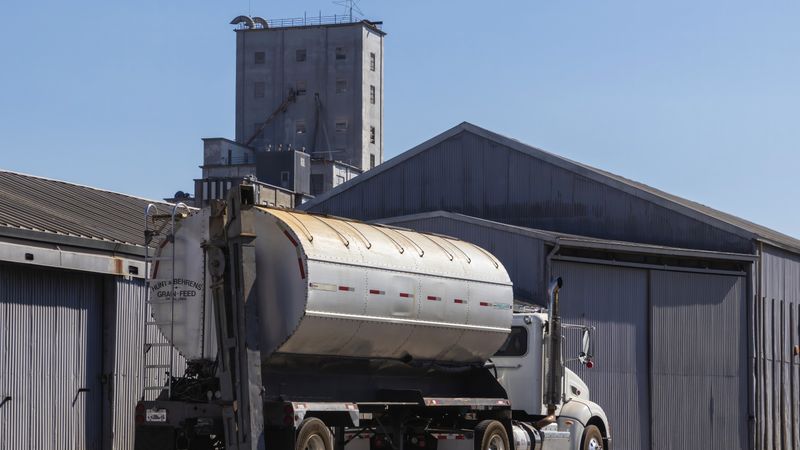 A large semi-trailer truck parked at an industrial grain warehouse under clear skies.