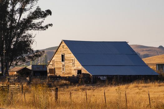 A wooden barn stands amidst a serene rural landscape, surrounded by fields and trees at dusk.