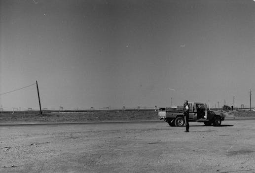 A black and white photograph of a man standing by a pickup truck in a desert landscape.