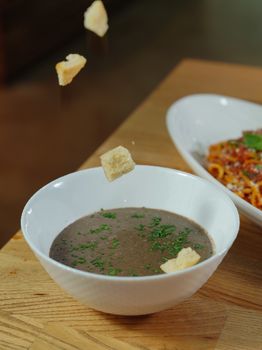 Mushroom soup in a white bowl with croutons falling, served on a wooden table.