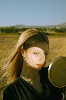 Stylish woman in a field holding a mirror, captured during sunrise with warm sunlight.