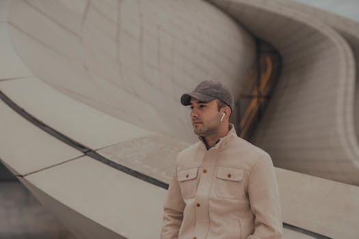 A stylish man wearing a cap and earphones stands against an architectural backdrop, embodying modern urban fashion.