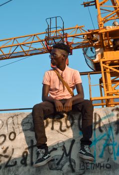 Young man in casual attire sitting against a construction site backdrop, showcasing urban style.