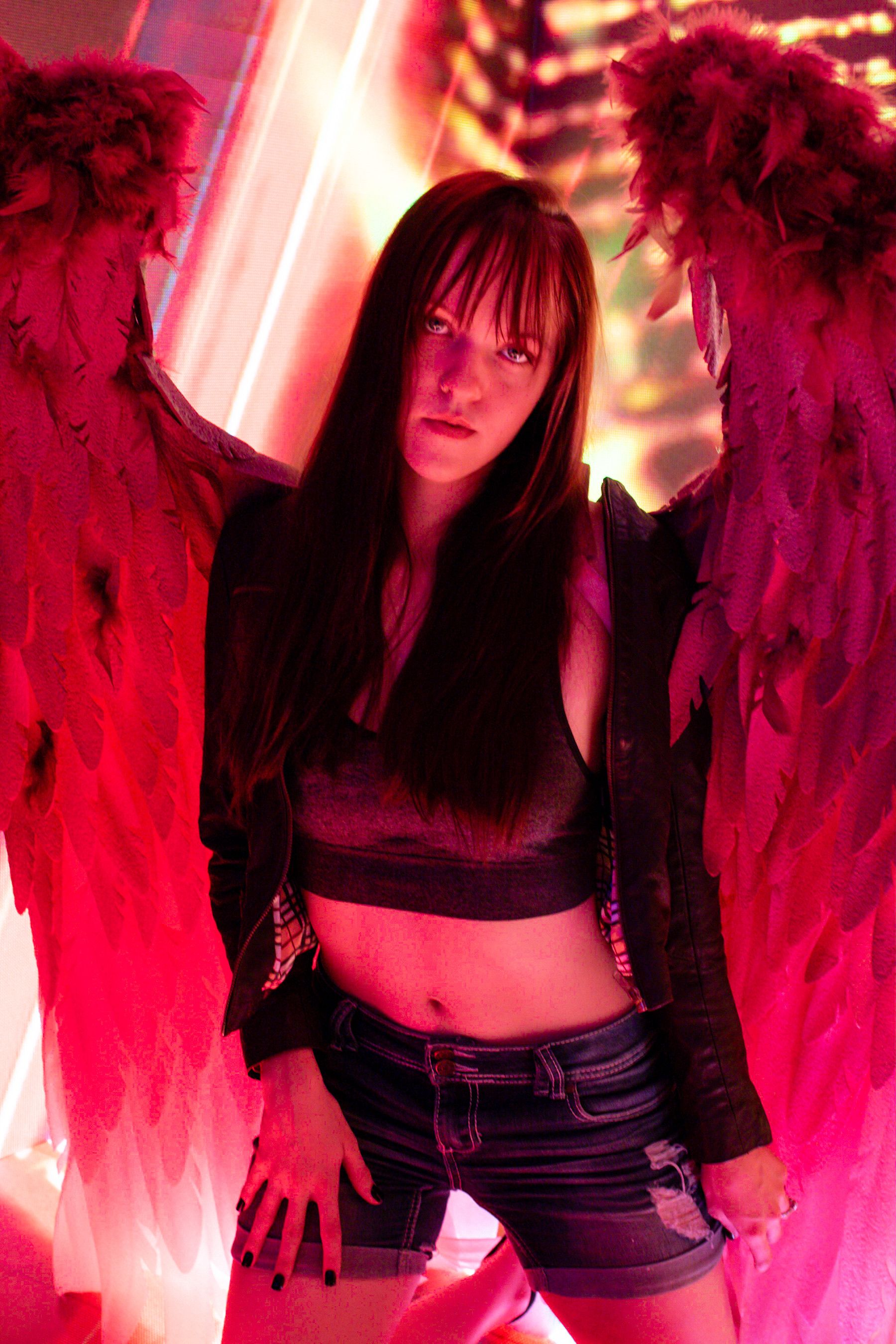 Model posing with angel wings in dramatic red lighting, fashion and fantasy fusion.