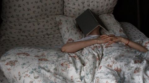 Woman relaxing in bed with book on face, floral bedding, creating a cozy vibe.