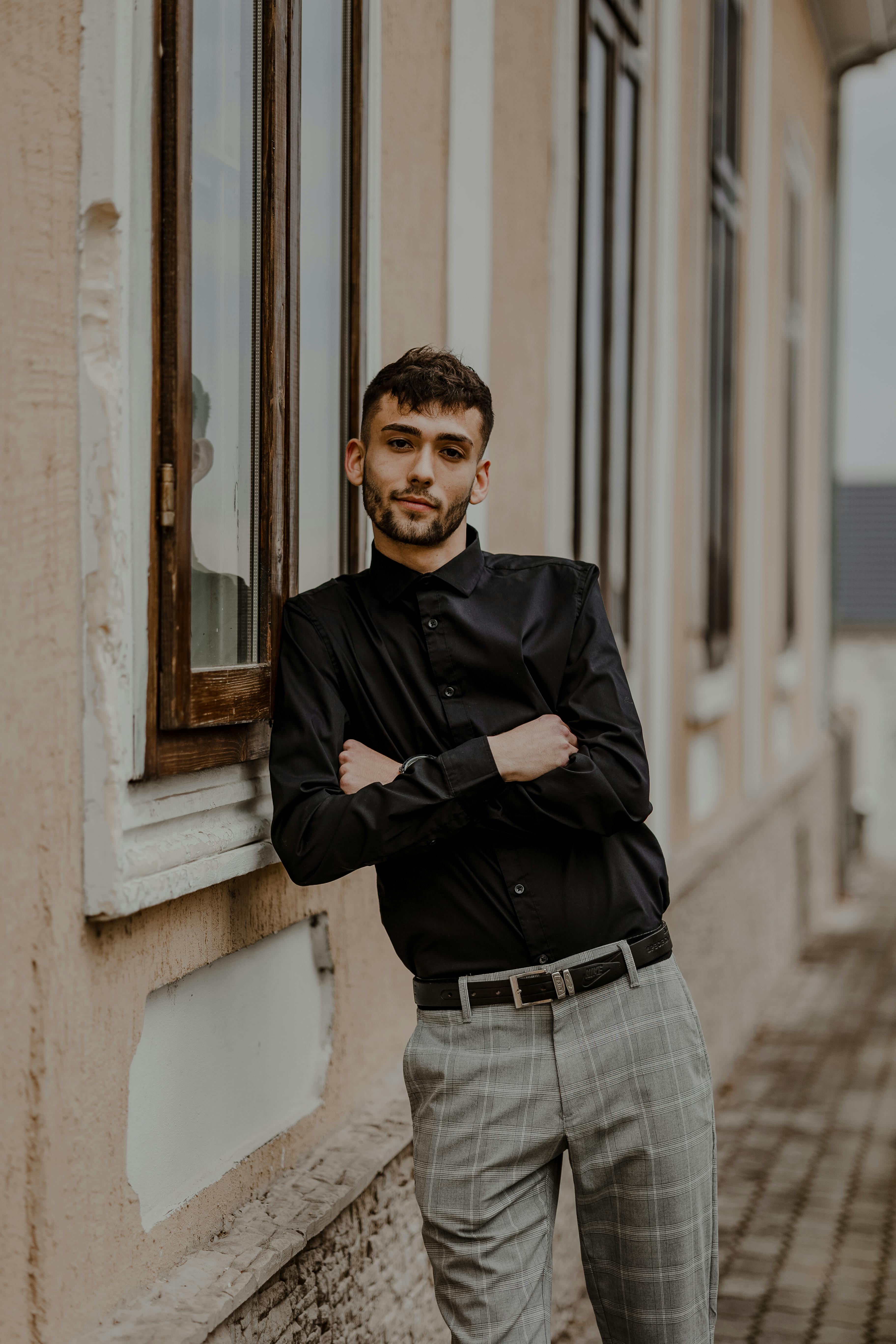 Fashionable young man in a black shirt poses with arms crossed in an urban street setting.