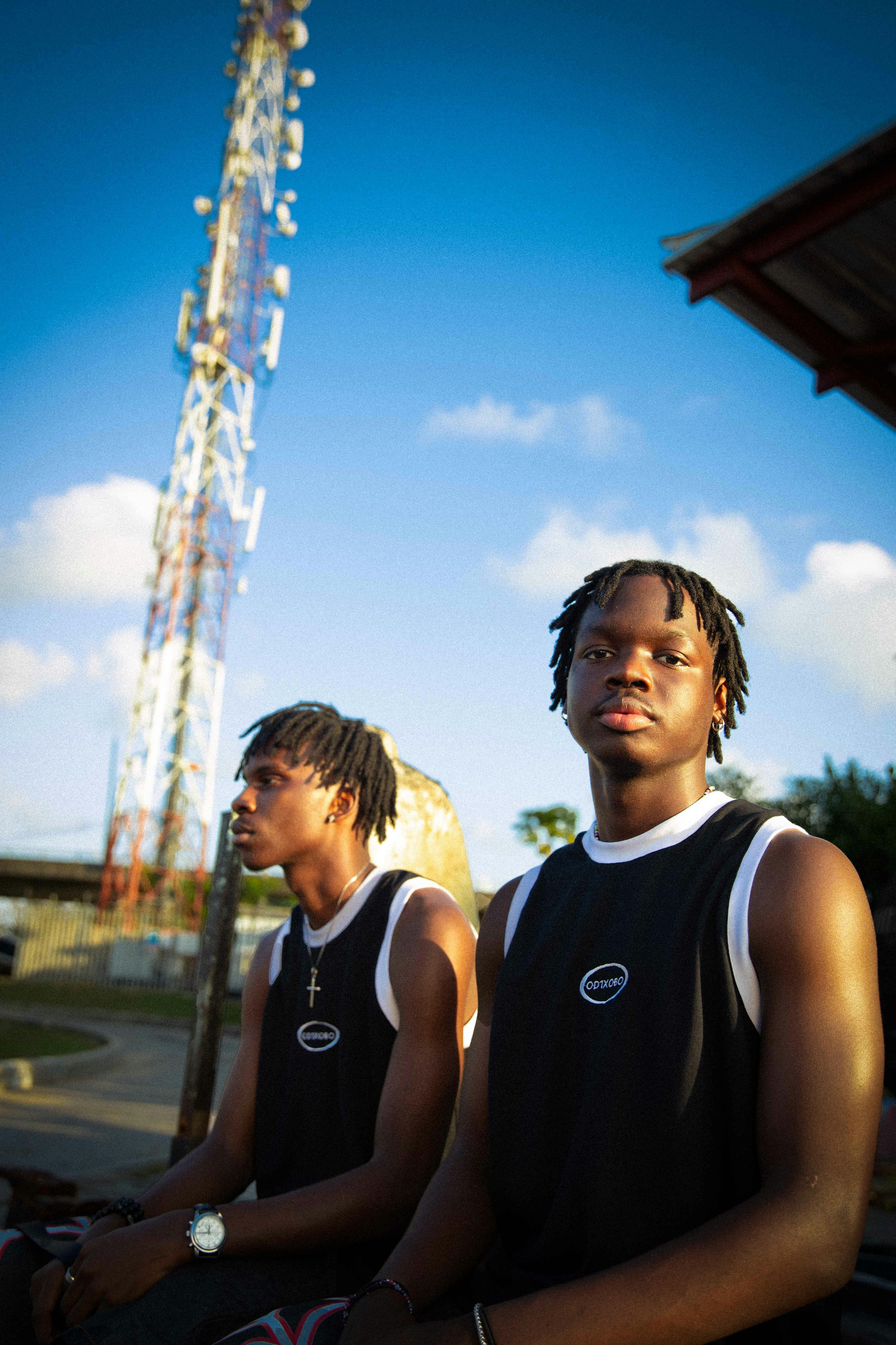 Two young men with dreadlocks pose outdoors against a bright sky, wearing sportswear and tank tops.