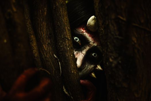 A person with devilish makeup peers through tree branches, creating an eerie Halloween vibe.