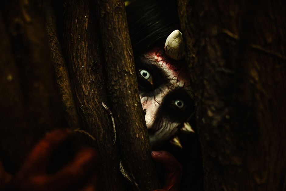 A person with devilish makeup peers through tree branches, creating an eerie Halloween vibe.