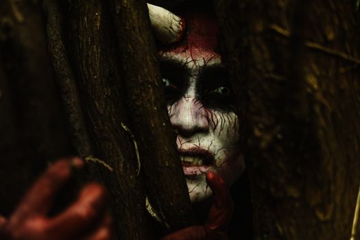 Scary demon-like face with stage makeup peeking through branches in a dark forest.