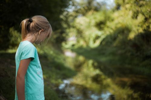Free A young girl stands thoughtfully by a tranquil stream in a lush park setting. Stock Photo