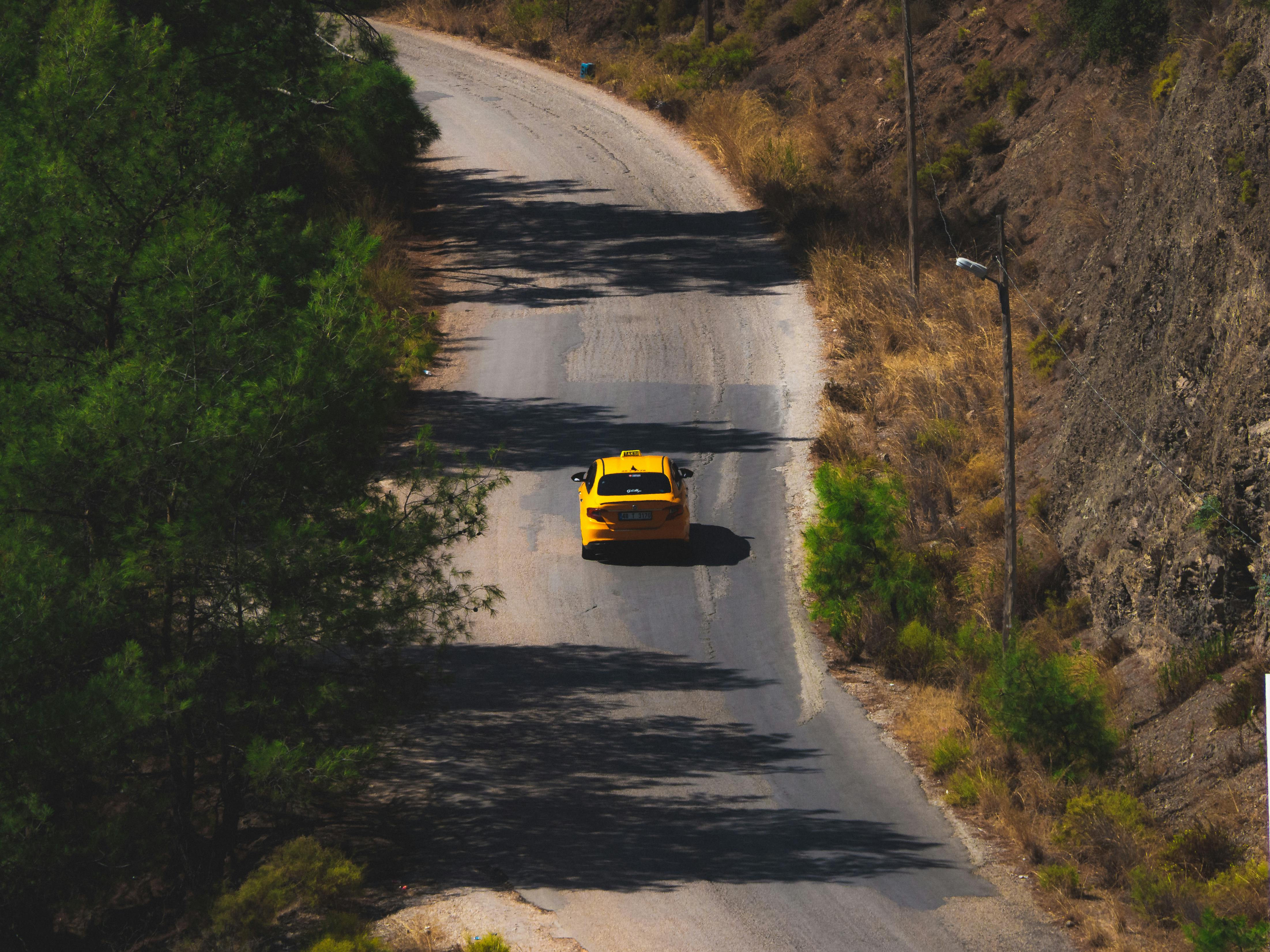 A yellow taxi travels along a winding road surrounded by countryside scenery.