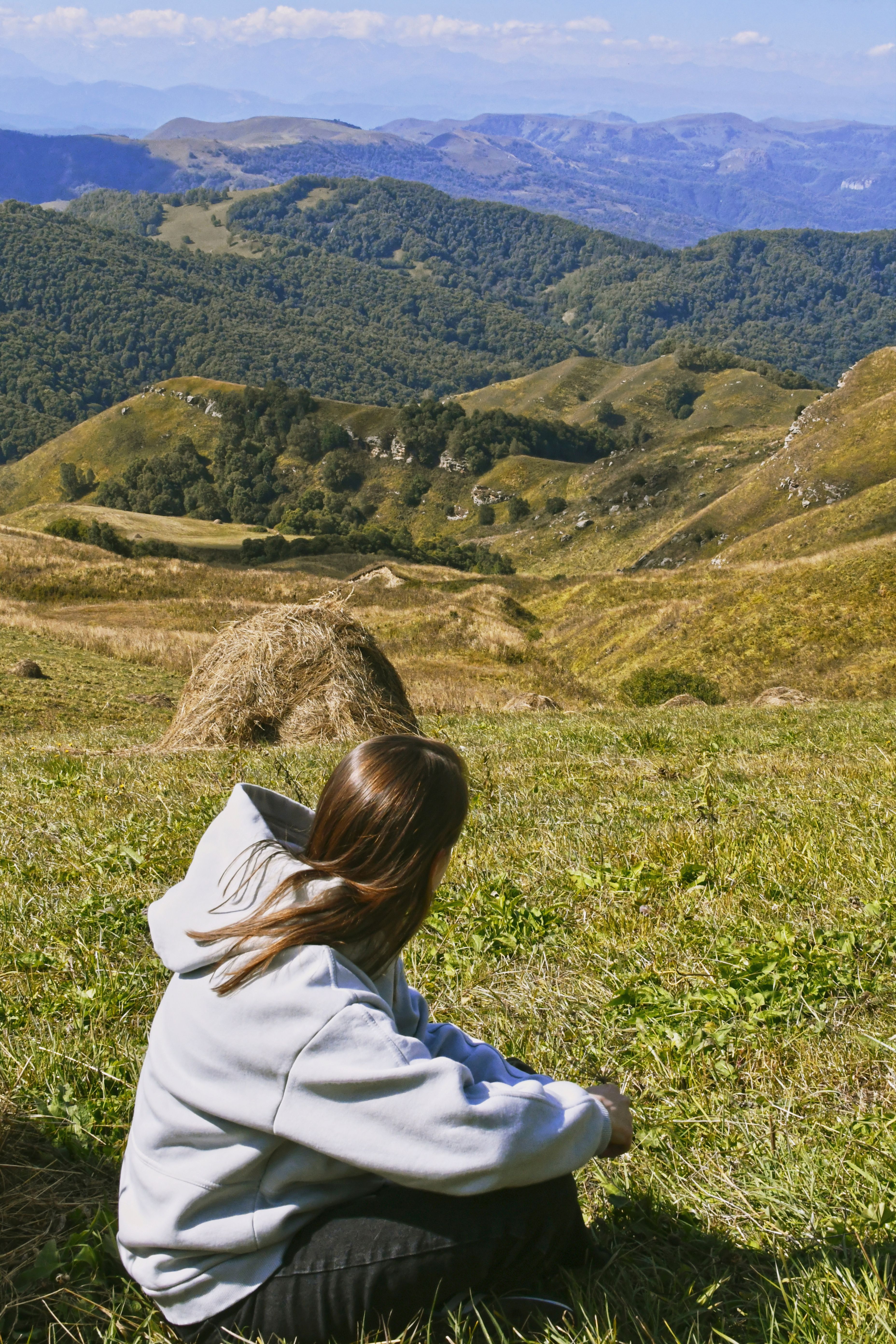 A woman in a hoodie sits in a scenic mountain landscape, enjoying nature.