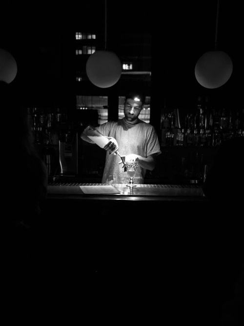 Free Black and white photo of a bartender working at night, pouring a drink in a dimly lit bar. Stock Photo