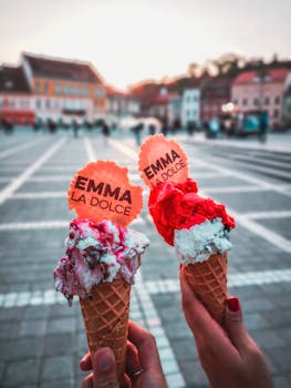 Two hands holding ice cream cones with colorful toppings in a scenic city square.