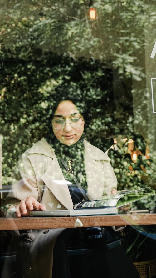 Free A woman in a headscarf reading behind a glass window, with green reflections creating a serene vibe. Stock Photo
