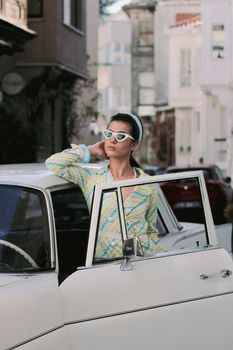 Chic woman in retro attire posing with a vintage car on a city street.