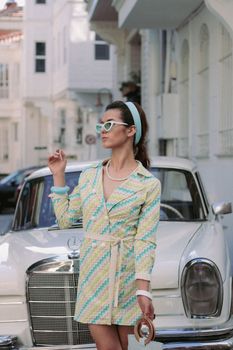 Stylish woman in retro attire posing by a classic vintage car on a sunny day.