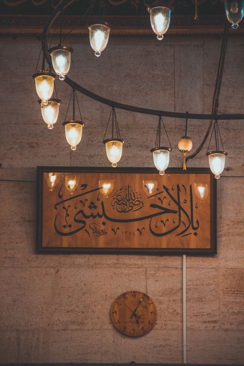 Free A serene mosque interior featuring Arabic calligraphy, a traditional chandelier, and clock. Stock Photo