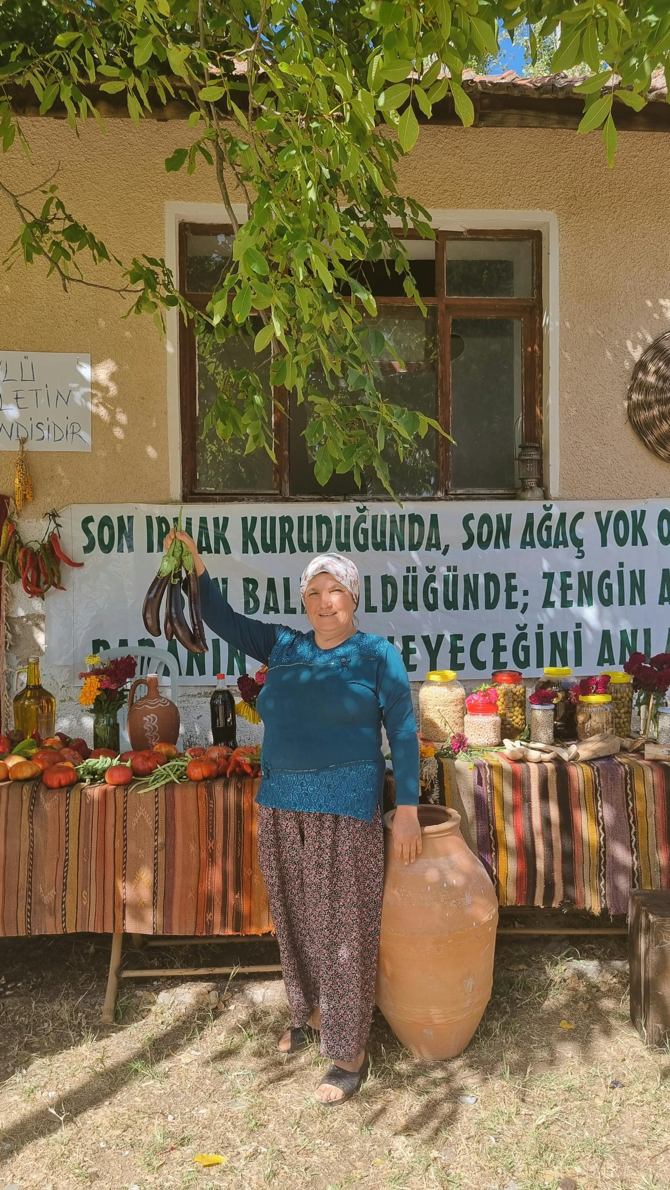 A woman stands proudly by a display of local produce and goods at a market in Deştin, Türkiye.