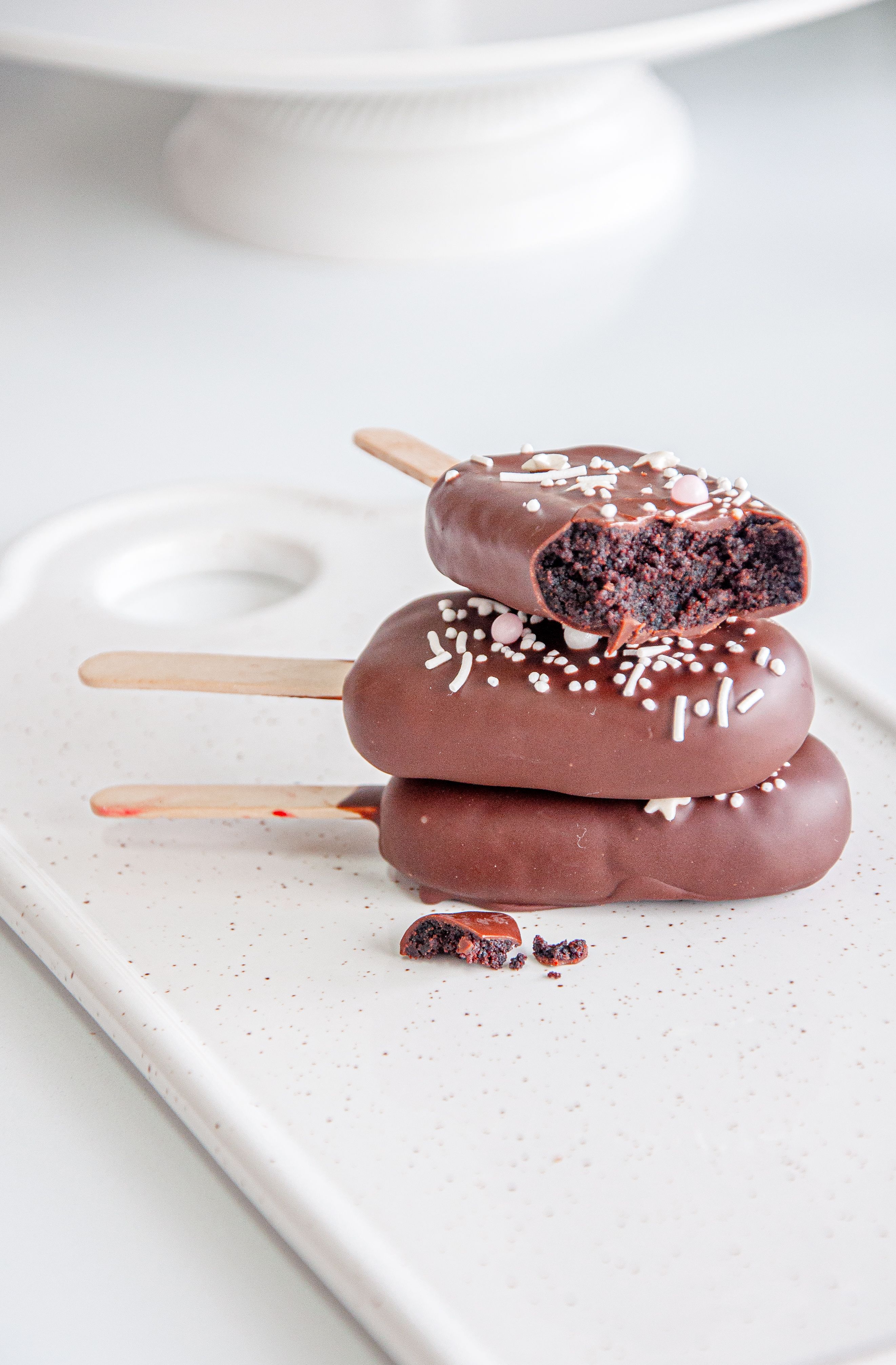 Popsicle-shaped cake pops (cakesicles) coated in pastel pink and white chocolate, decorated with gold flakes and delicate drizzles, displayed on a marble cake stand, fashion magazine styling