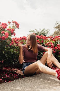 Young woman sitting among blooming red roses, enjoying a sunny day outdoors.