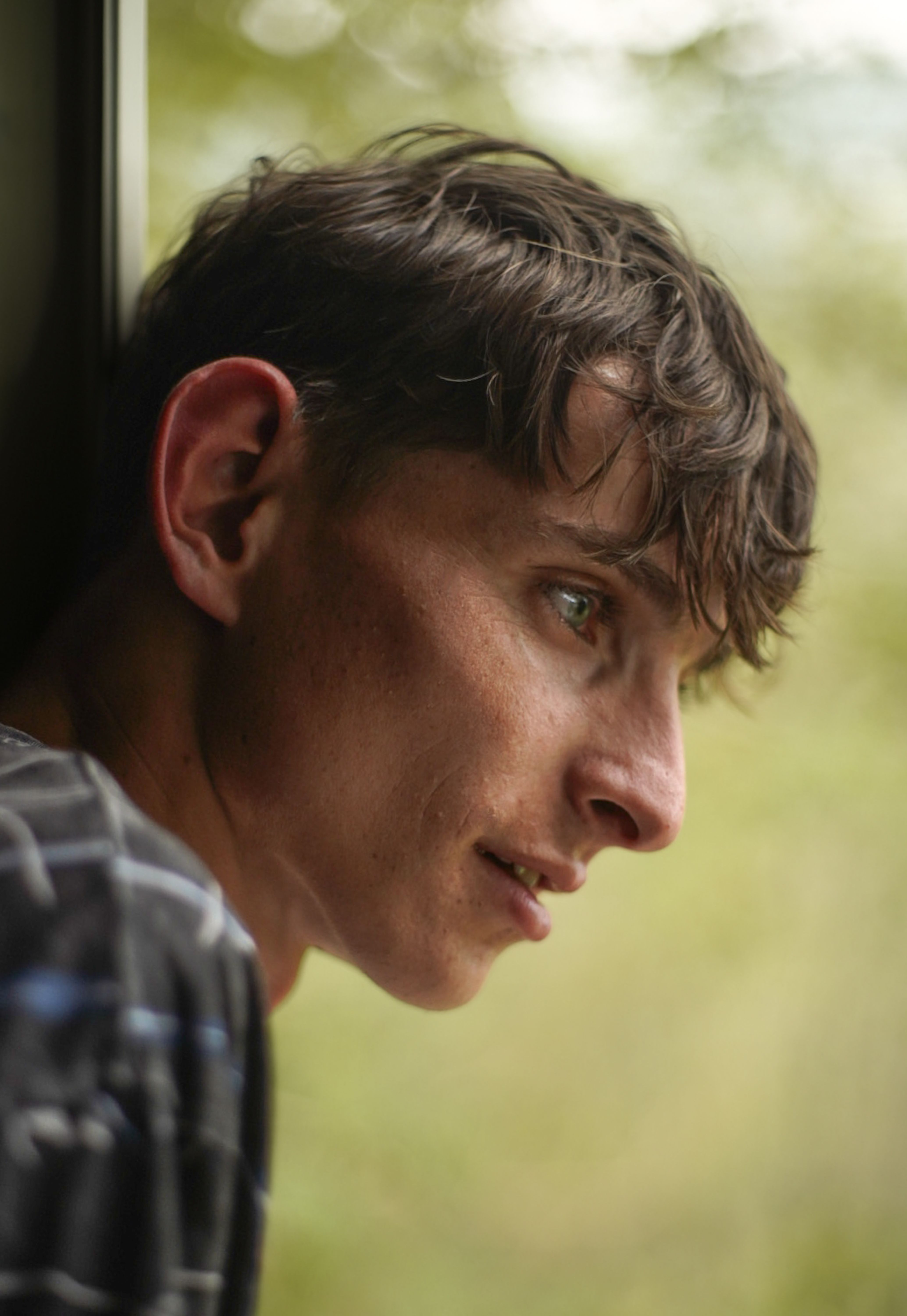 Young man with short brown hair leaning by window, deep in thought, surrounded by soft light.