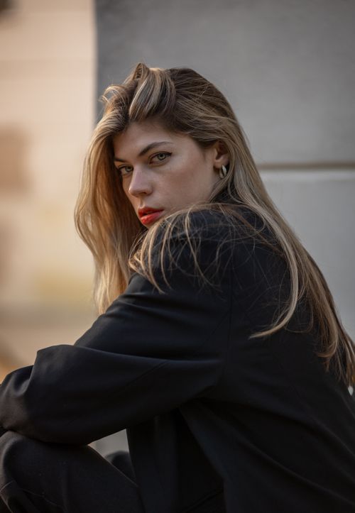 Free Elegant portrait of a blonde woman with long hair, wearing a black outfit, seated in an urban environment. Stock Photo