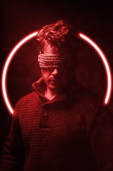 A man blindfolded, surrounded by red light and smoke, creating a dramatic atmosphere.