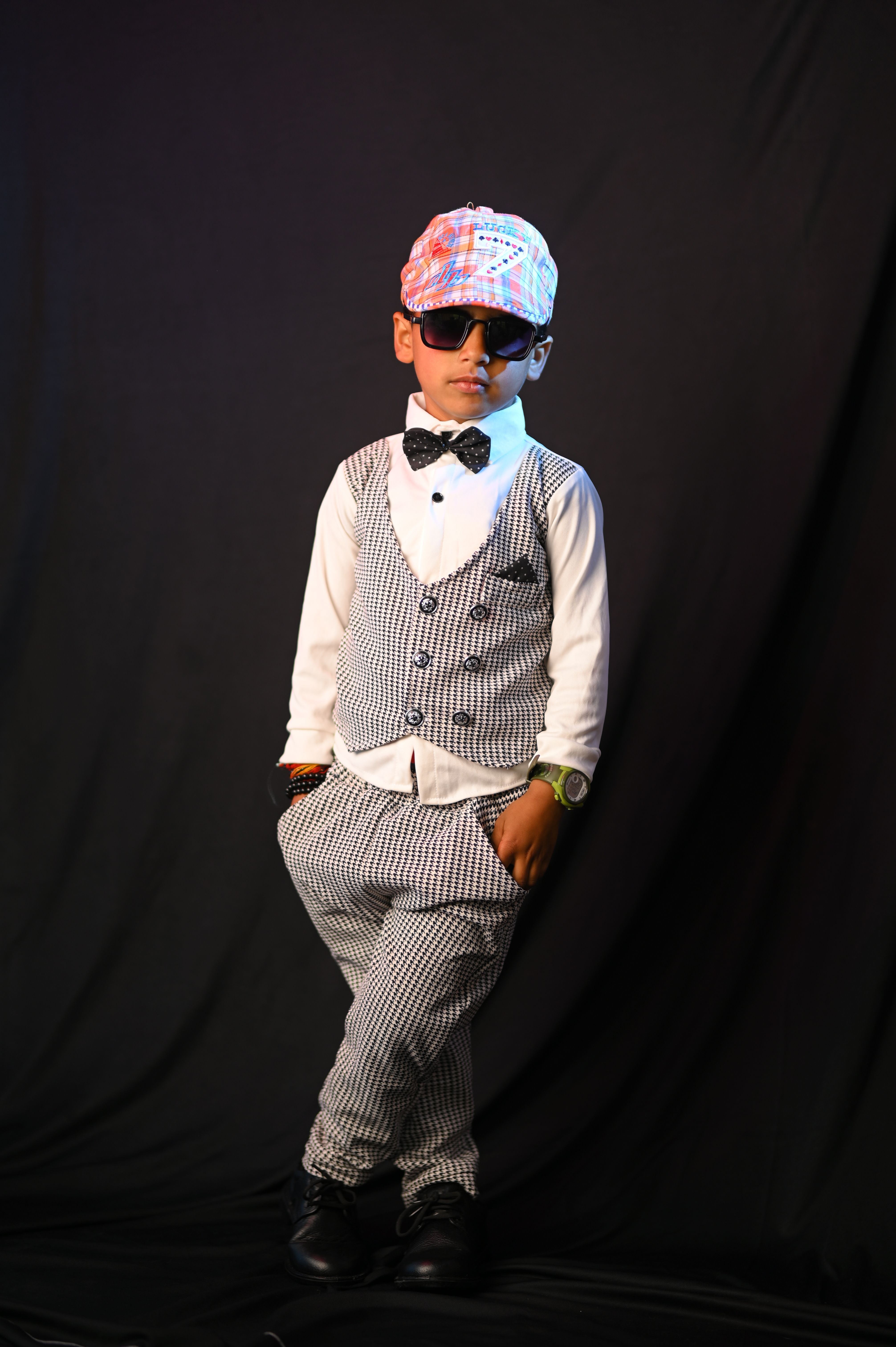 A young boy striking a pose in a stylish fashion outfit with a cap and sunglasses against a dark background.