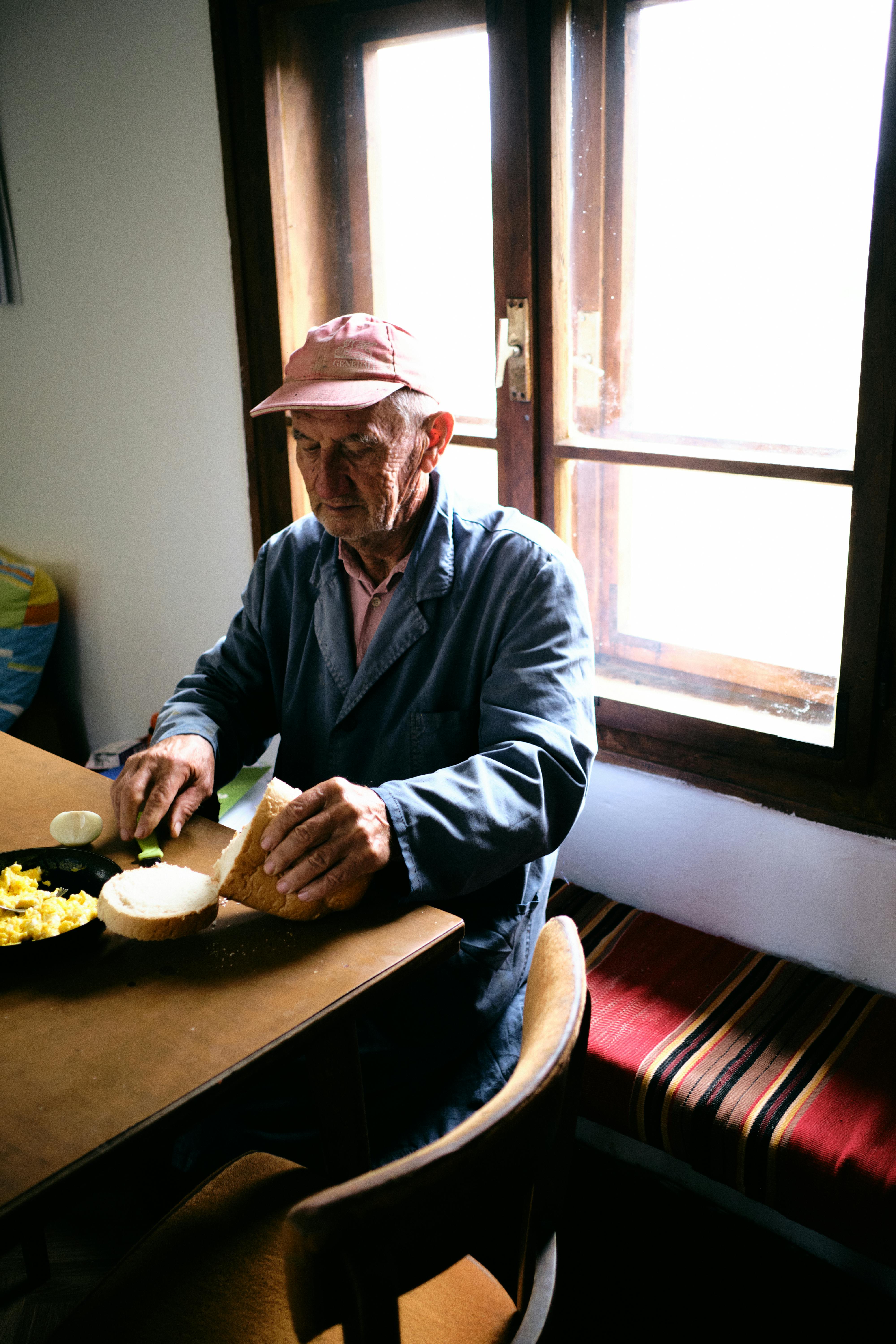 Elderly man in cap sitting at table for breakfast by the window. Cozy natural light setting.