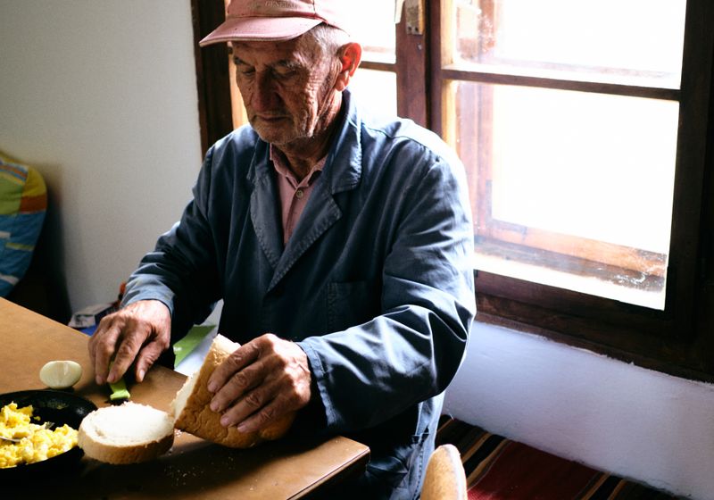 An older man in a blue work jacket slices a loaf of white bread at a wooden table beside a bright window, a pan of scrambled eggs in front of him