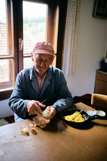 Elderly man peeling onions by window while preparing breakfast indoors.