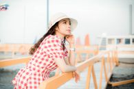 一位头戴帽子、身穿红色方格连衣裙的妇女，正在码头上享受阳光明媚的夏日，尽显悠闲