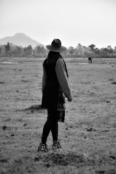 Black and white photo of a woman in a hat standing in a grassy field facing a distant mountain.