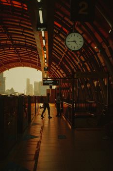 Silhouetted passengers walking on a platform in a Jakarta train station, under an arched roof and a large clock.