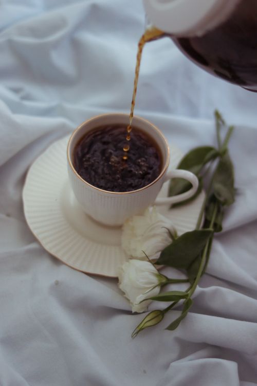Free Hot coffee being poured into a cup beside white roses on a textured fabric backdrop. Stock Photo