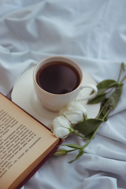 Free Cup of coffee on a white bed next to an open book and flowers, creating a cozy morning vibe. Stock Photo