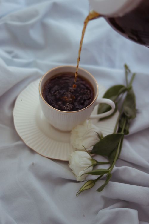 Free A close-up of coffee being poured into a white cup next to flowers on a bed. Stock Photo