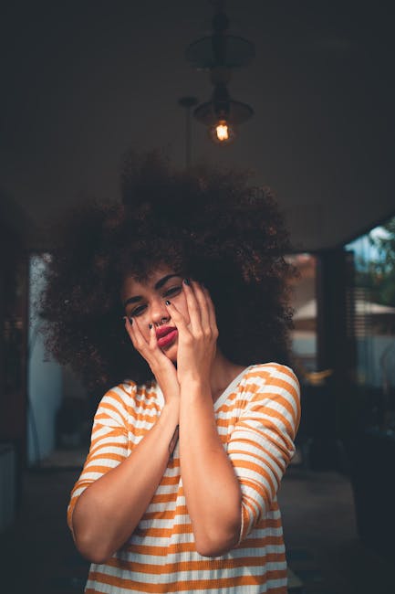 A woman with afro hair posing indoors, expressing thoughtfulness in casual attire.